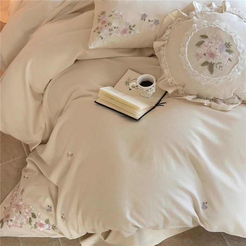 Beige bedding set with floral embroidery on a bed, featuring a book and cup.