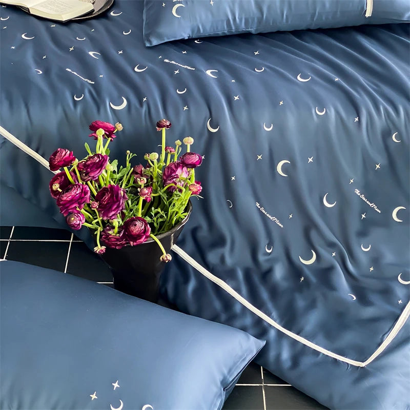 Blue bedding set with moon and star pattern, accompanied by a vase of purple flowers.