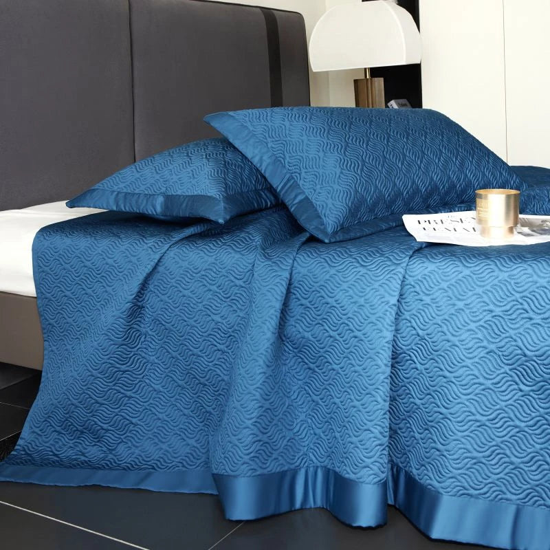 Blue textured bedspread on a bed with pillows and a lamp in the background.