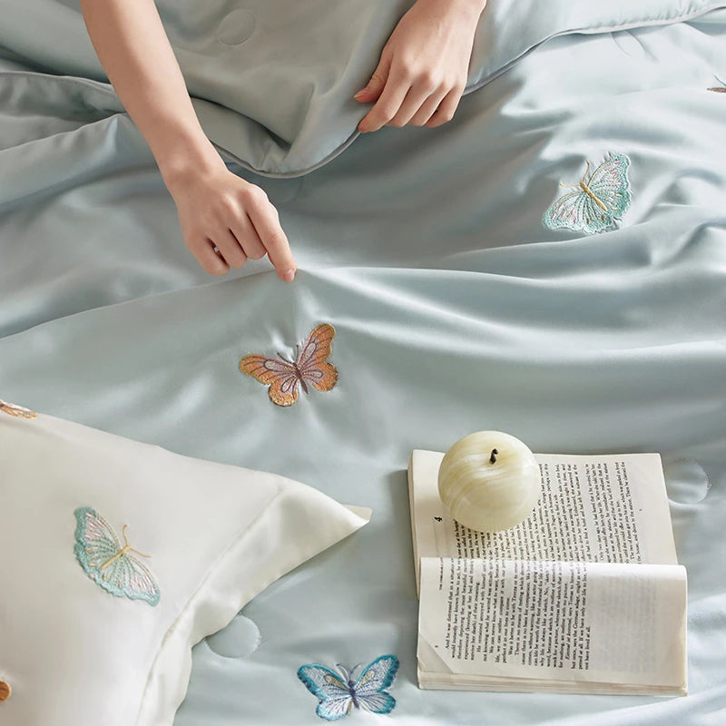 Embroidered butterfly patterns on a light blue bedding set with a hand and book in the foreground.