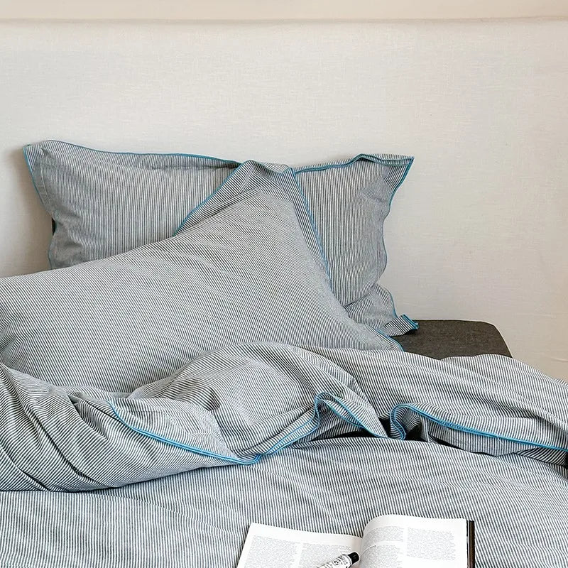 Gray striped bedding with blue piping on a bed against a white wall.