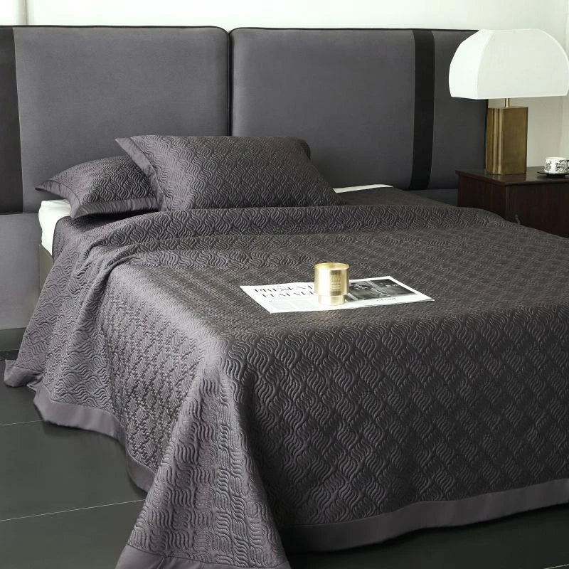 Gray textured bedspread on a bed with a nightstand and lamp in a bedroom setting.