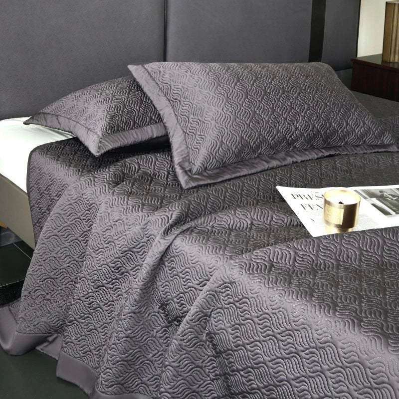 Gray textured bedspread on a bed with pillows and a lamp in the background