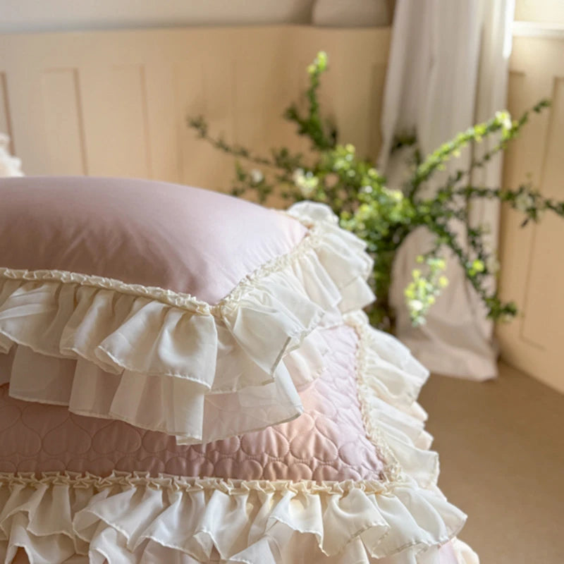 Pink ruffled pillow with lace trim on a bed in a softly lit room.
