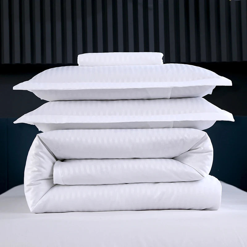Stack of white bedding including duvet, comforter, and pillows on a dark background