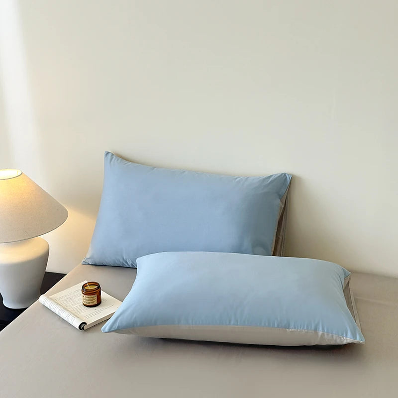 Two light blue pillows on a bed next to a lamp and small table with a book and jar.