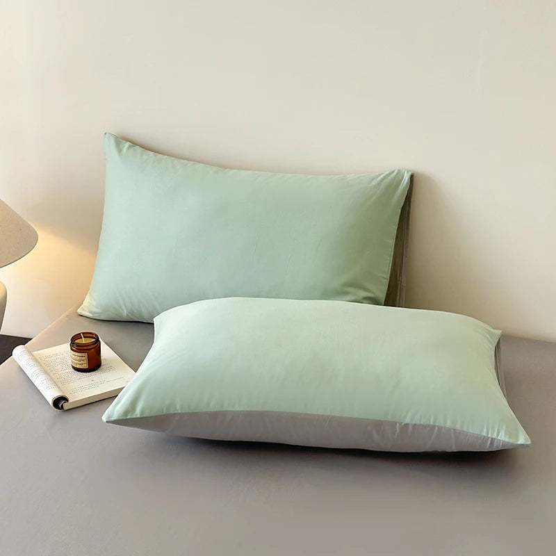 Two light green pillows on a bed with a candle and book.