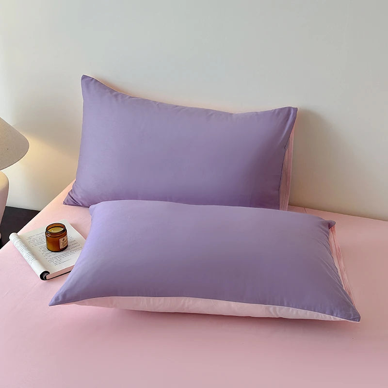 Two purple pillows on a pink bed with a book and candle.