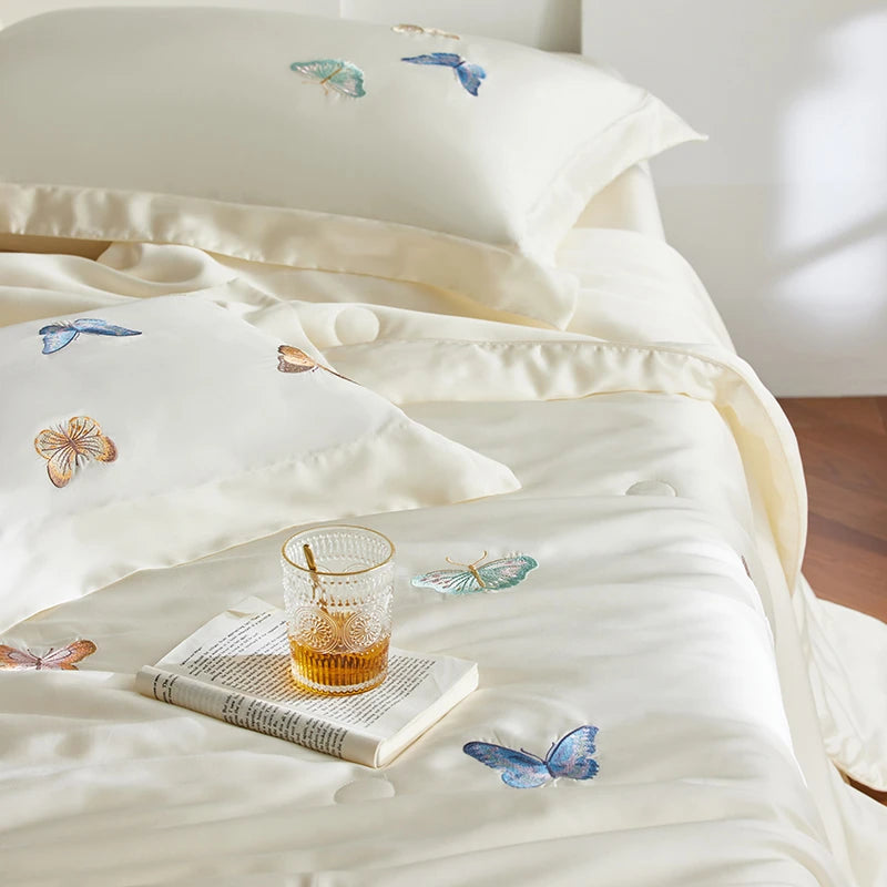 White bedding set with butterfly patterns on a bed, accompanied by a glass of amber liquid and a book.